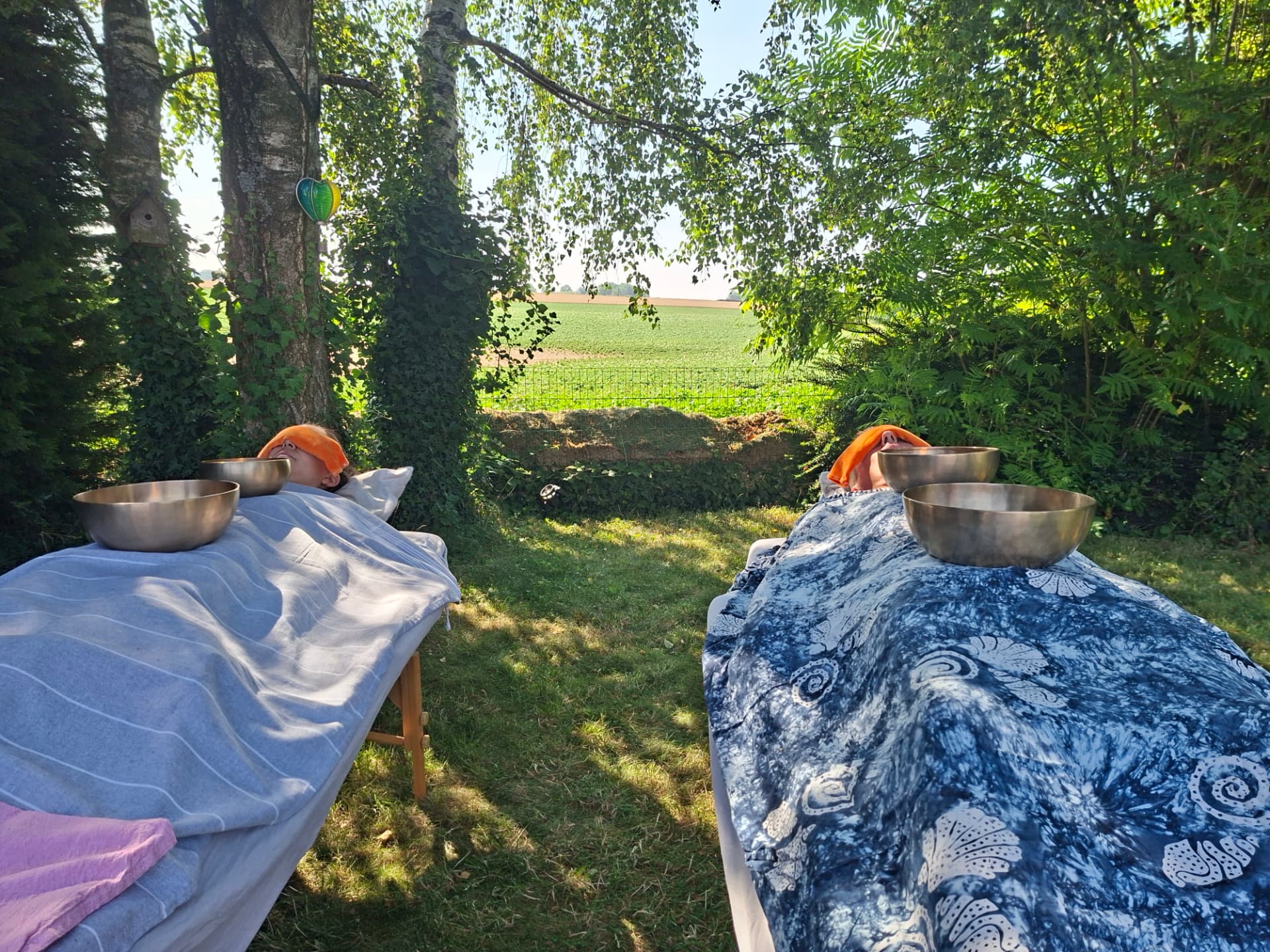 Massage sonore en duo en extérieur : deux personnes allongées sous des plaids, bols chantants posés sur elles, face à la campagne